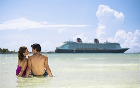 A man and a woman in bathing suits sit on the beach with a Disney Cruise Line ship in the background