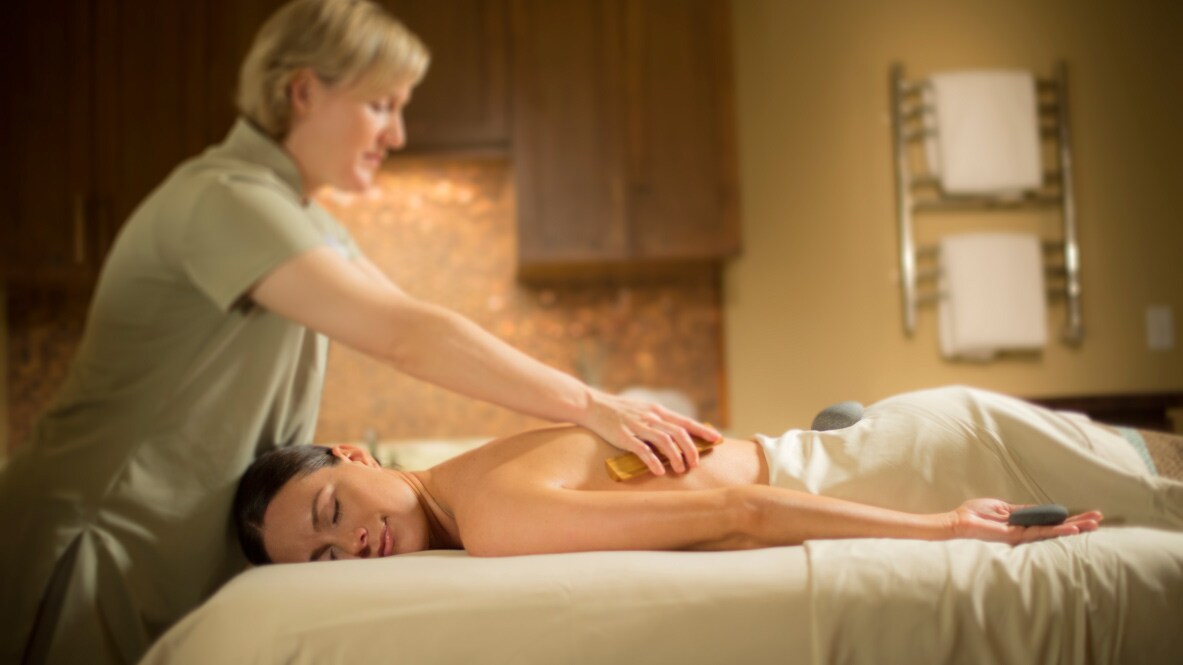 A woman lies face-down on a massage table, while a masseuse massages her shoulders